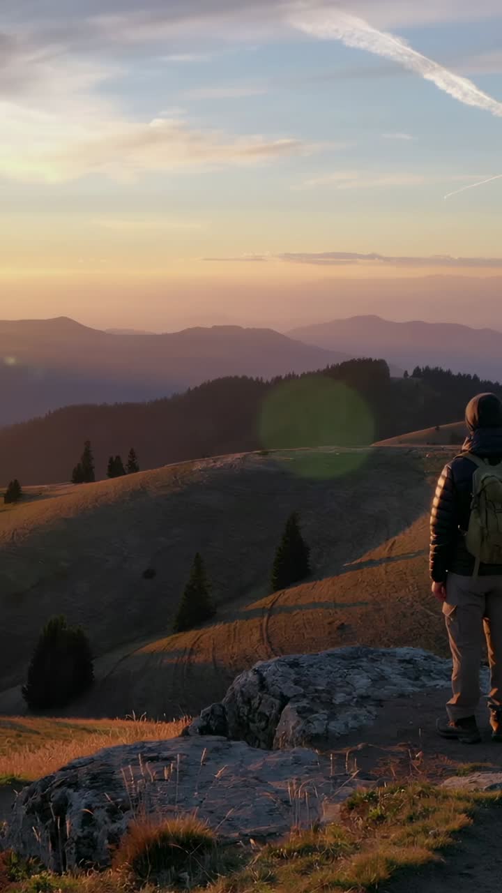 Vertical video: Sunset drawing hiker on ledge watching horizon in puffy coat, pack, copy space