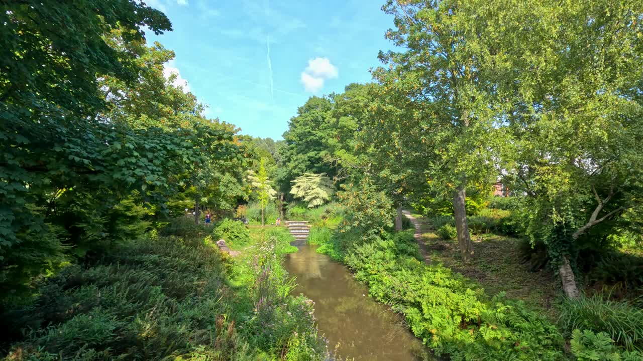 Wide shot of lush green canal, trees, and walkway under bright daylight, static camera