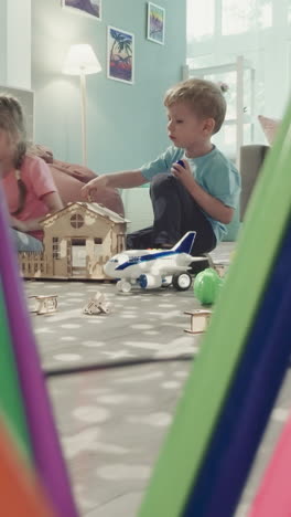 siblings decorate roof of wooden house with colorful plastic blocks. girl and boy play with toys in nursery view through clearance in bunch of pencils in glass