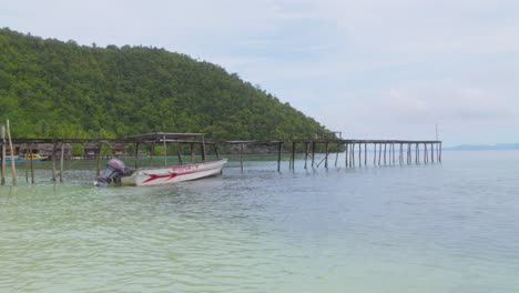 a wooden pier extending into the calm turquoise waters, with a motorboat anchored nearby and lush green hills in the background