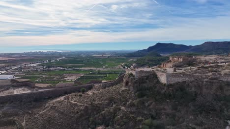 lateral-flight-with-parallax-effect-in-a-castle-discovering-its-dimension-and-in-the-background-we-see-fields-of-orange-crops-with-mountains,-a-town-and-the-sea,-there-is-a-blue-sky-with-clouds