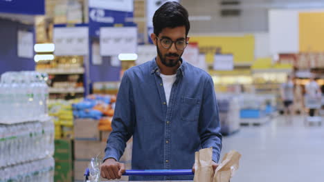 portrait of a tall boy with glasses leaning on the shopping cart in the supermarket looking straight at the camera and smiling