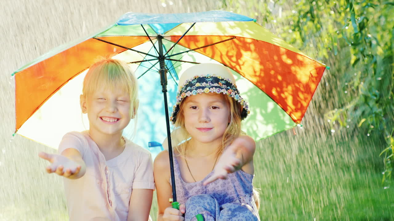 두 명의 평온한 소녀가 따뜻한 여름 비를 피해 우산 아래에 앉아 있습니다.
