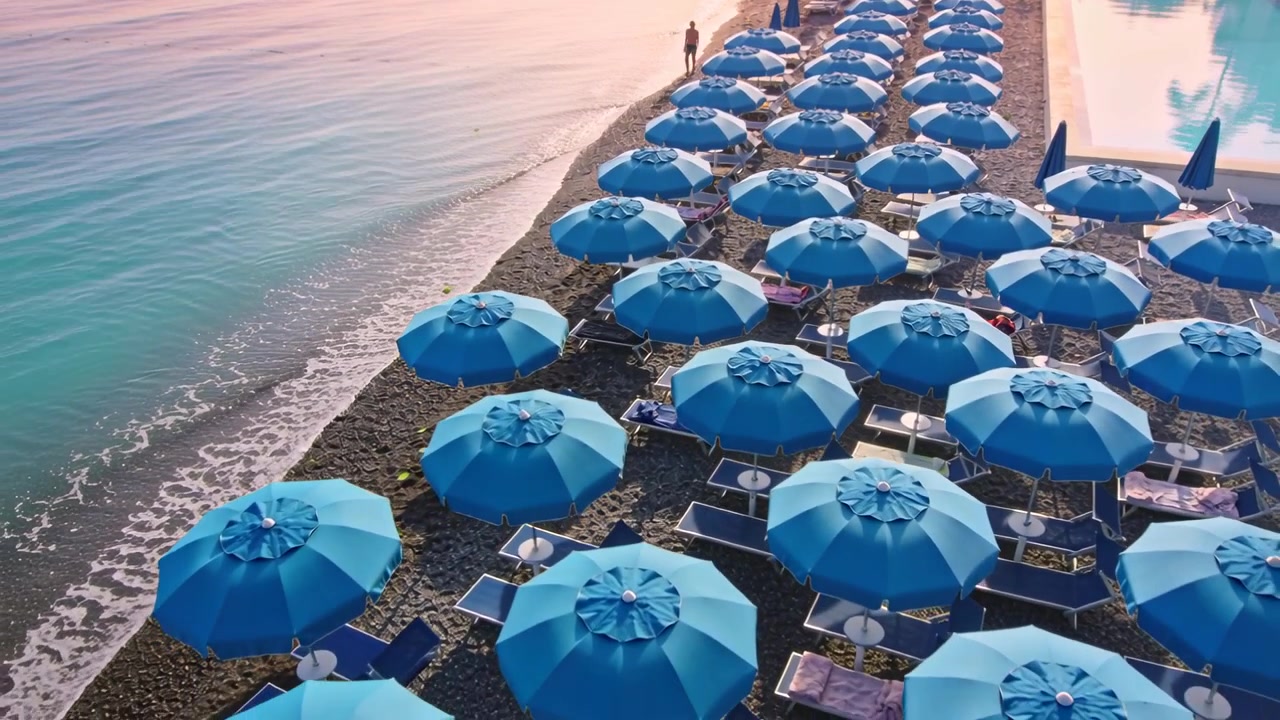 Drone che sorvola e traccia file di ombrelloni e un uomo in costume da bagno che cammina lungo la riva catturando l'alba.