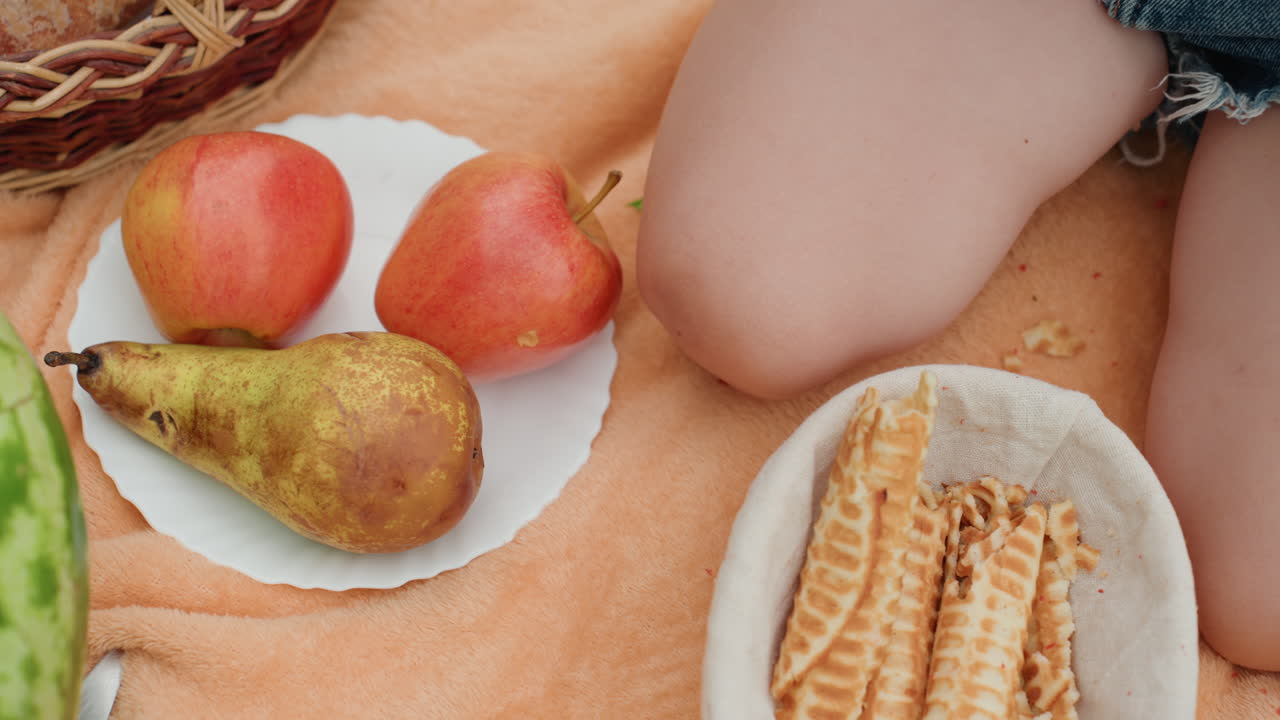 melancia e peras em toalha com perna vestida com jeans visível, prato de frutas maduras e biscoitos wafer em manta laranja, cesto de vime na borda, cena de lazer ao ar livre iluminada pelo sol, evocando lanches casuais
