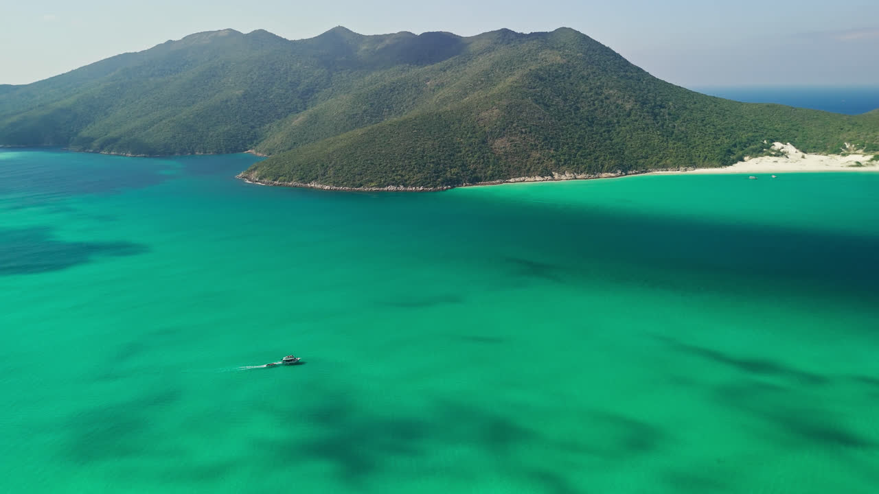 드론이 아레알 도 카보 (arraial do cabo) 의 순수한 파란색 해안 해역을 가로질러 날아다니며 배경에는 웅장한 섬 산과 함께 운행하는 배를 포착합니다.
