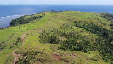 Footpath-trail-on-gentle-green-slopes-of-a-hilly-island,-Aerial-seascape