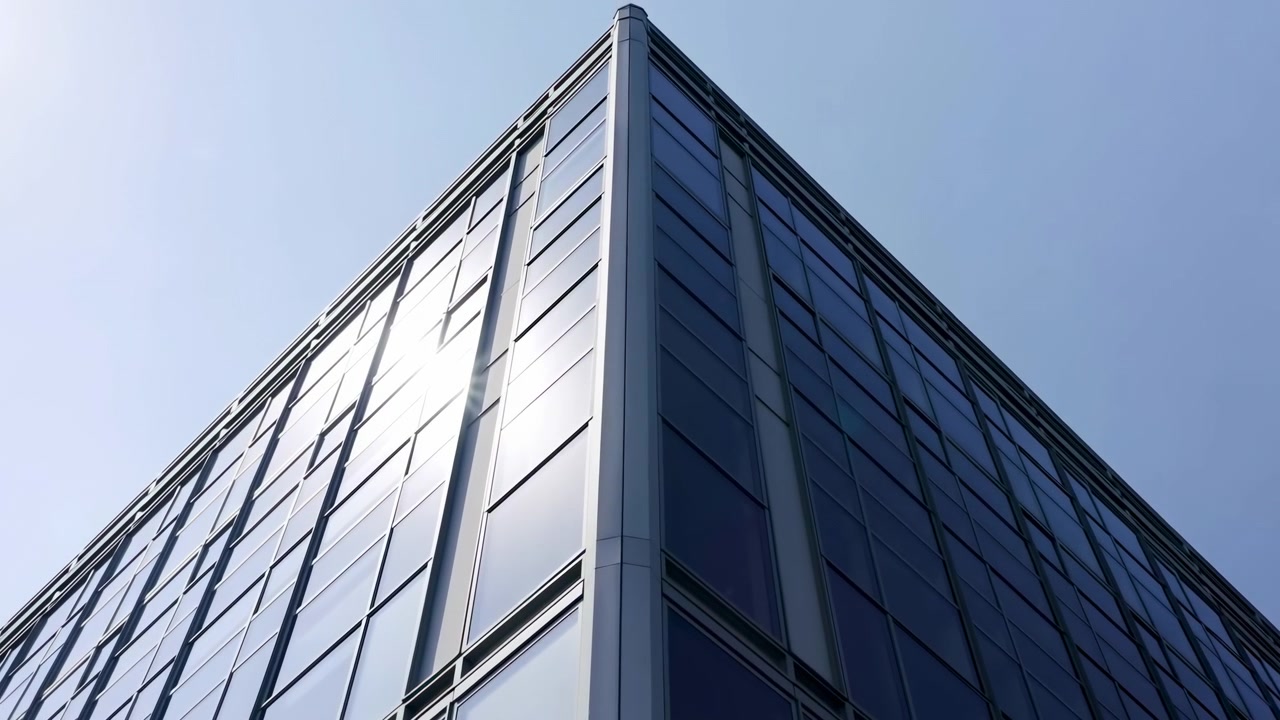 La vista se abre gradualmente alrededor de la esquina de un edificio prominente, mientras la luz del sol brilla a través de paneles de vidrio tintados de azul, exponiendo más líneas de la fachada, superficies reflectantes, molduras metálicas y el cielo diurno abierto
