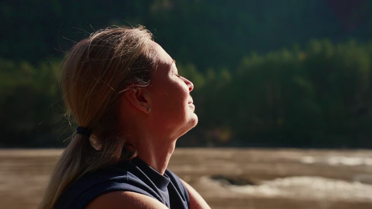 Eine Frau mit blondem Haar blickt mit geschlossenen Augen nach oben und lächelt friedlich. Sie steht nahe einem ruhigen Fluss mit bewaldeten Hügeln im Hintergrund. Sonnenlicht streichelt sanft ihr Gesicht und erzeugt warme, sanfte Glanzlichter.