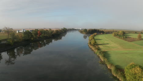 serene calm morning with early autumn color on trees seen from drone flying over calm river flowing through farm fields