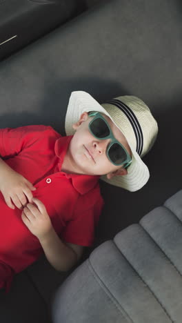 sweet toddler in straw hat puts hands on head back lying on rotating sofa. little boy in sunglasses with green rim relaxes on yacht upper close view