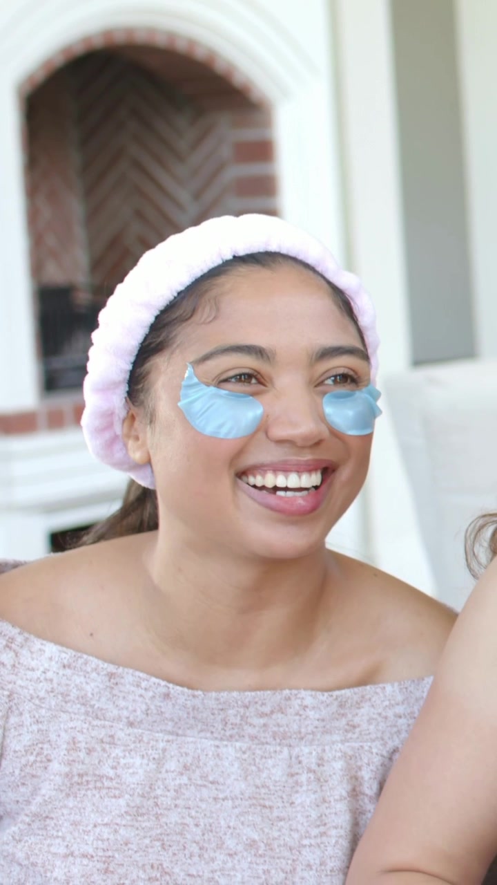 Vertical video: Smiling friend, left woman laughing on sofa with pink headband and blue eye patches