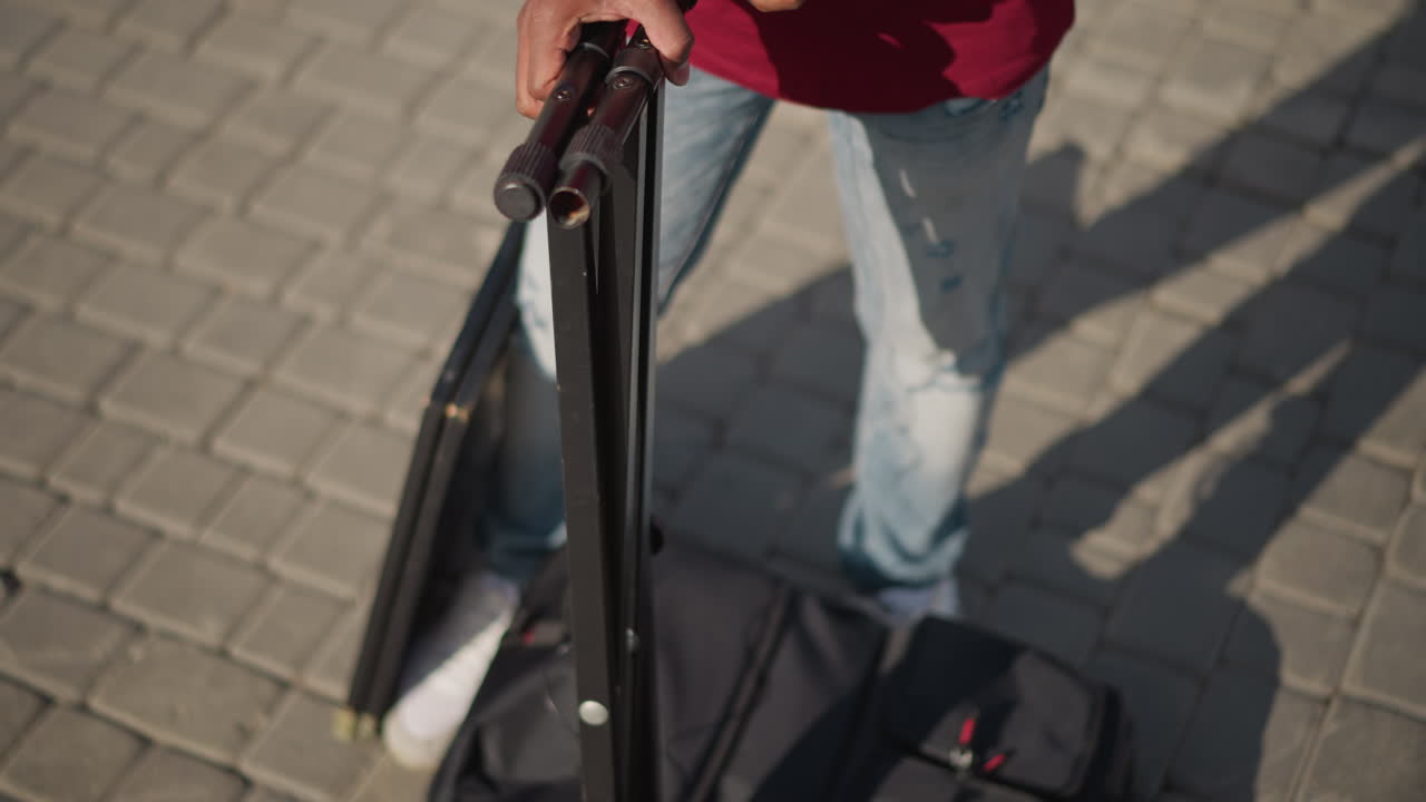 Nahaufnahme von Händen, die einen Ständer auf dem Bürgersteig aufbauen. Eine Draufsicht zeigt den Künstler, wie er Klammer und Gelenk festzieht, Finger greifen ein Metallstativ neben einem Rucksack auf einem Kopfsteinpflasterplatz. Eine sorgfältige Routine des Befestigens und Anpassens.