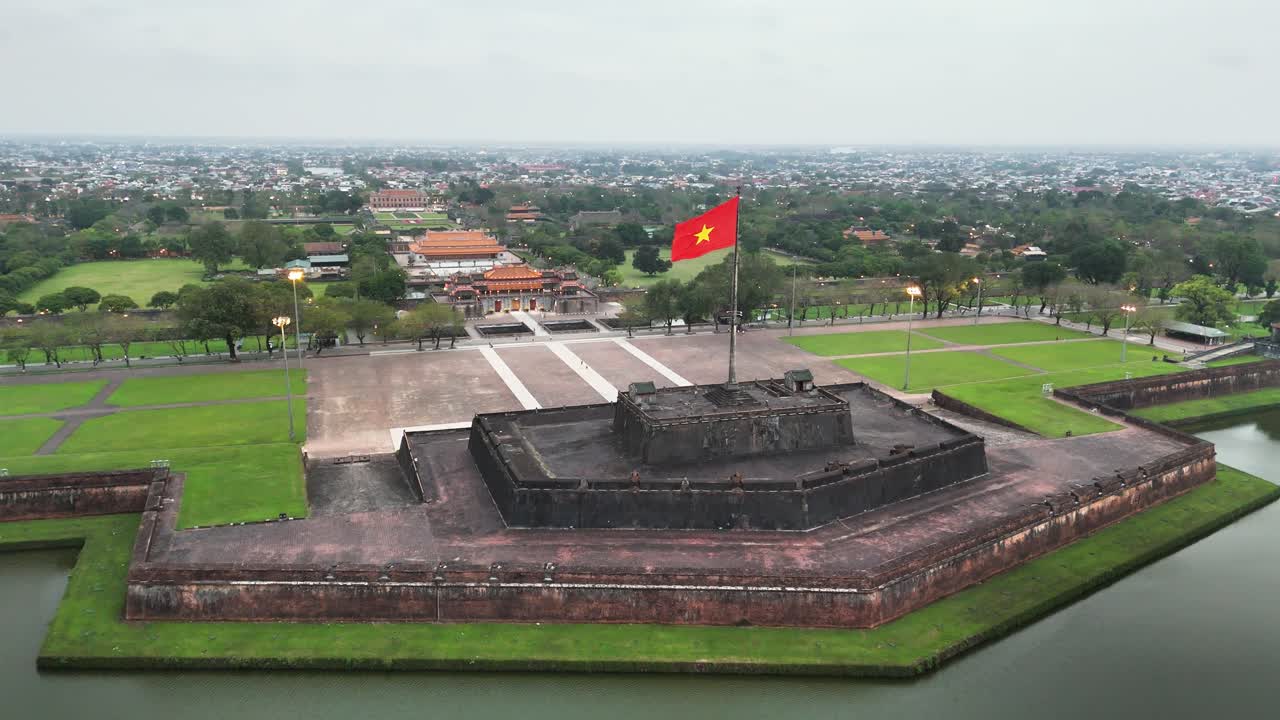 Historic flag tower at Hue Citadel standing above stone walls and moat, with imperial palace complex beyond, on the banks of the Perfume River, central Vietnam, captured by elevated drone shot push in