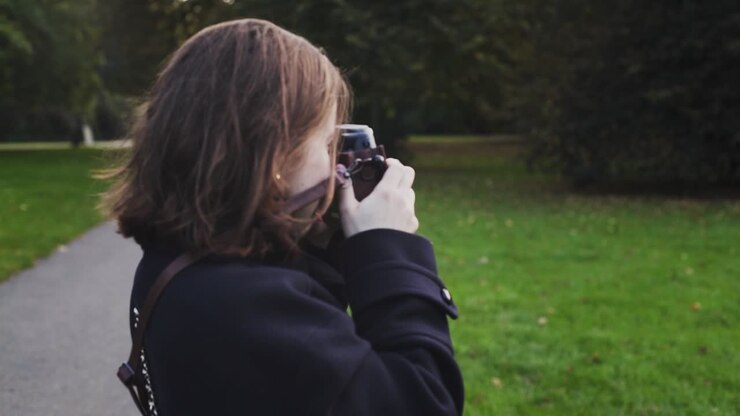 Young Caucasian brunette woman wearing jacket takes photograph outside by green grass in outdoor park with camera, close up profile slow motion