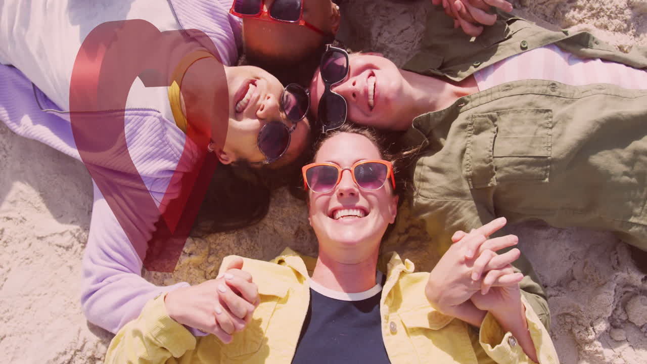 Wearing sunglasses, diverse friends lying on sand holding hands, smiling together