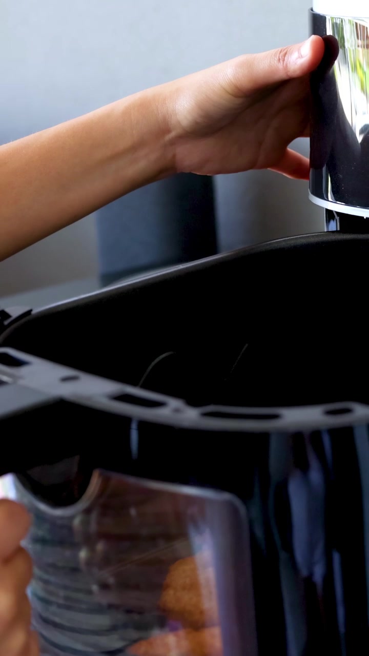 Mani che azionano una friggitrice ad aria, rimuovendo cibo cotto in un ambiente cucina luminoso. Il video cattura il processo con illuminazione chiara e naturale