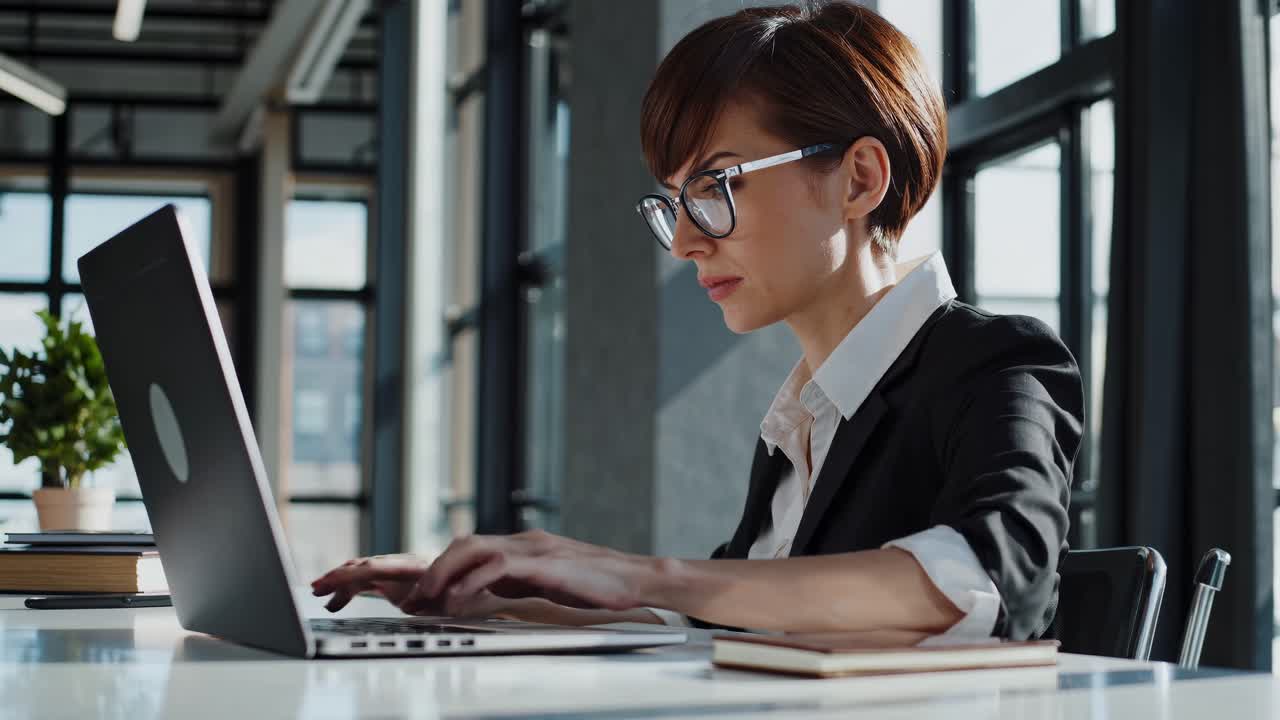 A professional woman in glasses works on a laptop in a modern office
