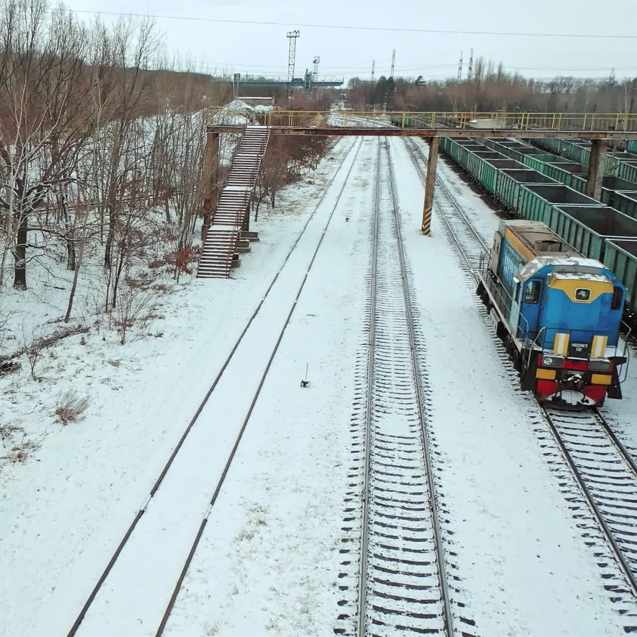 A freight train is passing by rail along rows of containers outside the city in the station area in the winter. Aerial view.