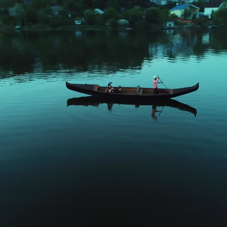 aerial view of a strong man in hat rowing with the oar in a boat over the river background at night. woman enjoys the beautiful landscape in the evening sitting in gondola.