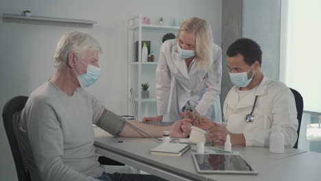 a young black doctor in a facemask takes blood pressure to a grey haired man under the gaze of a female blonde doctor