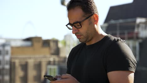 A-person-stands-outdoors-on-a-sunny-day,-engrossed-in-their-mobile-phone