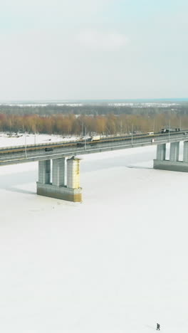 cars drive along long grey bridge over frozen river with walking person against forest on sunny day upper view