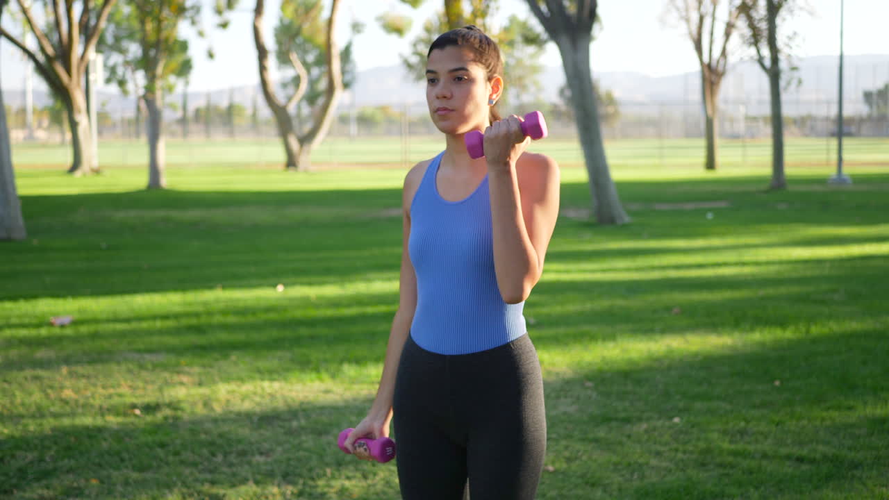 A young woman doing a dumbbell workout in the park performing bicep curls to build arm muscle