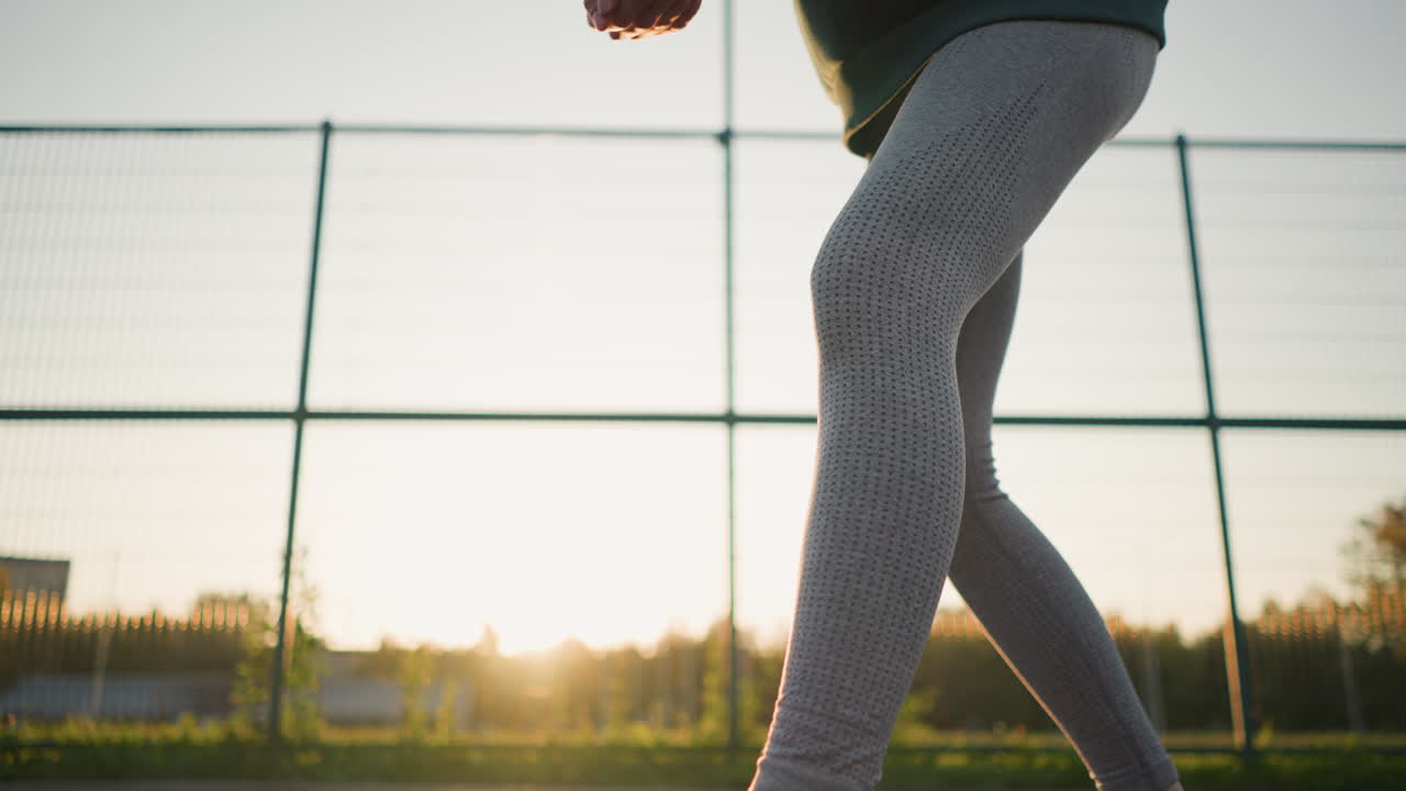 스포츠 드레스를 입은 여성이 풀, 울타리, 빛으로 배경에 골든 아워 동안 배구를 하는 낮은 각도 시각, 역동적인 야외 스포츠 분위기를 만니다.