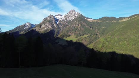 Person-Mit-Schwarzer-Jacke-Sitzt-Im-Schatten-Neben-Dem-Gipfelkreuz-Mit-Berglandschaft-Im-Hintergrund-Mit-Schneebedecktem-Gipfel-An-Einem-Sonnigen-Tag-In-Österreich,-Europa