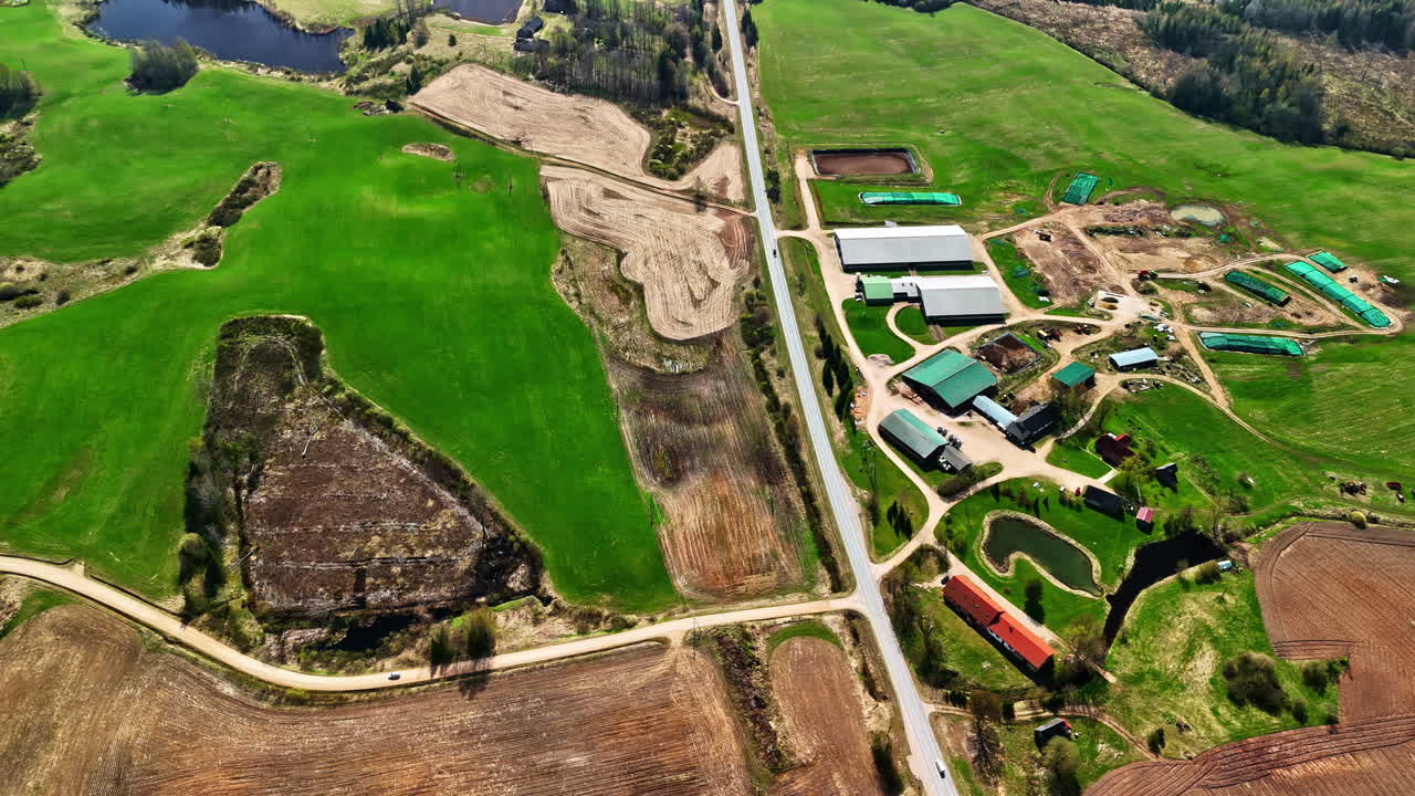 Aerial drone view of a modern farm complex with barns and houses next to a road surrounded by green fields and plowed land in rural Latvia
