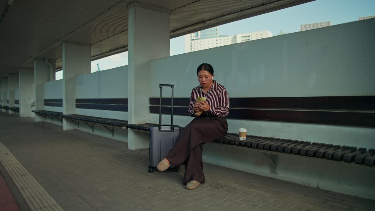 Mujer sentada en un banco con equipaje, comiendo un sándwich y bebiendo café