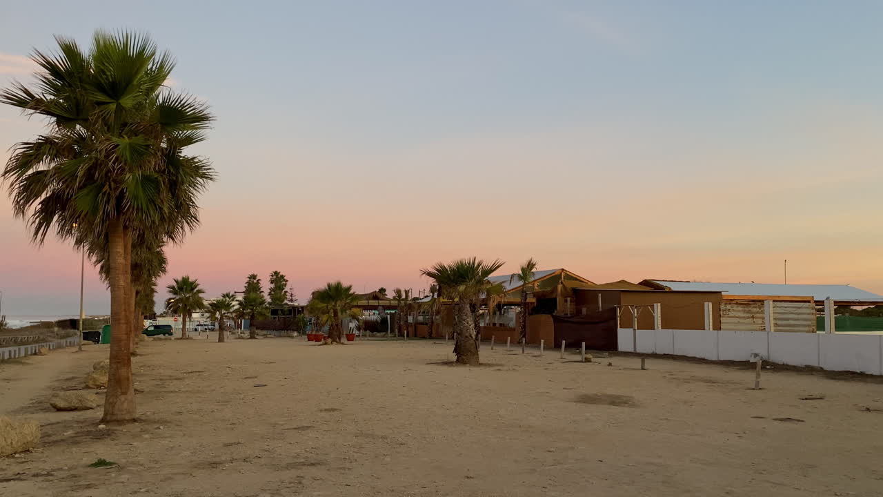 Beach bars and palms at El Palmar glow under a soft pastel sky, waiting for the next summer of salty nights and surf stories