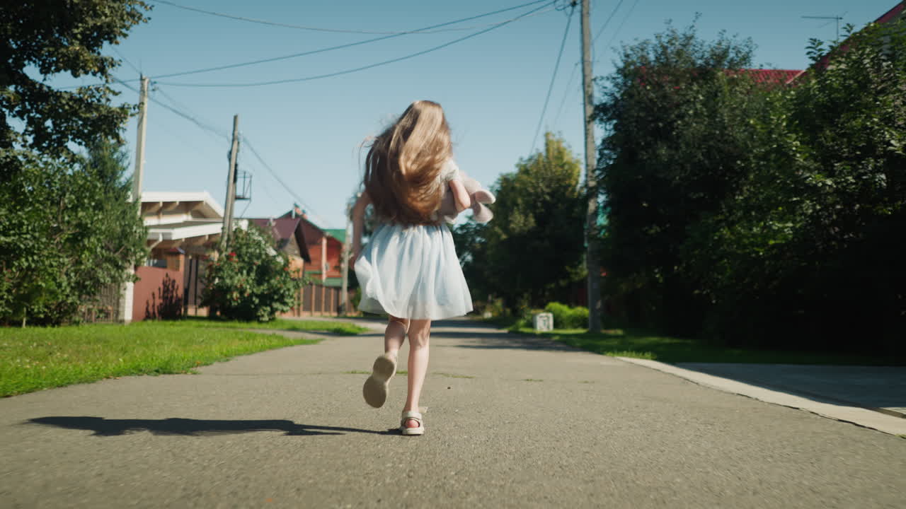 Back view of little girl in light dress running along sunny suburban street with hair bouncing in motion and toy in hand, surrounded by houses, trees, and power lines under clear sky