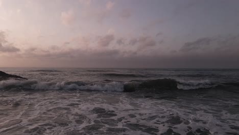 low angle aerial pull back across dramatic sunset rocky splashing coastal beach skyline