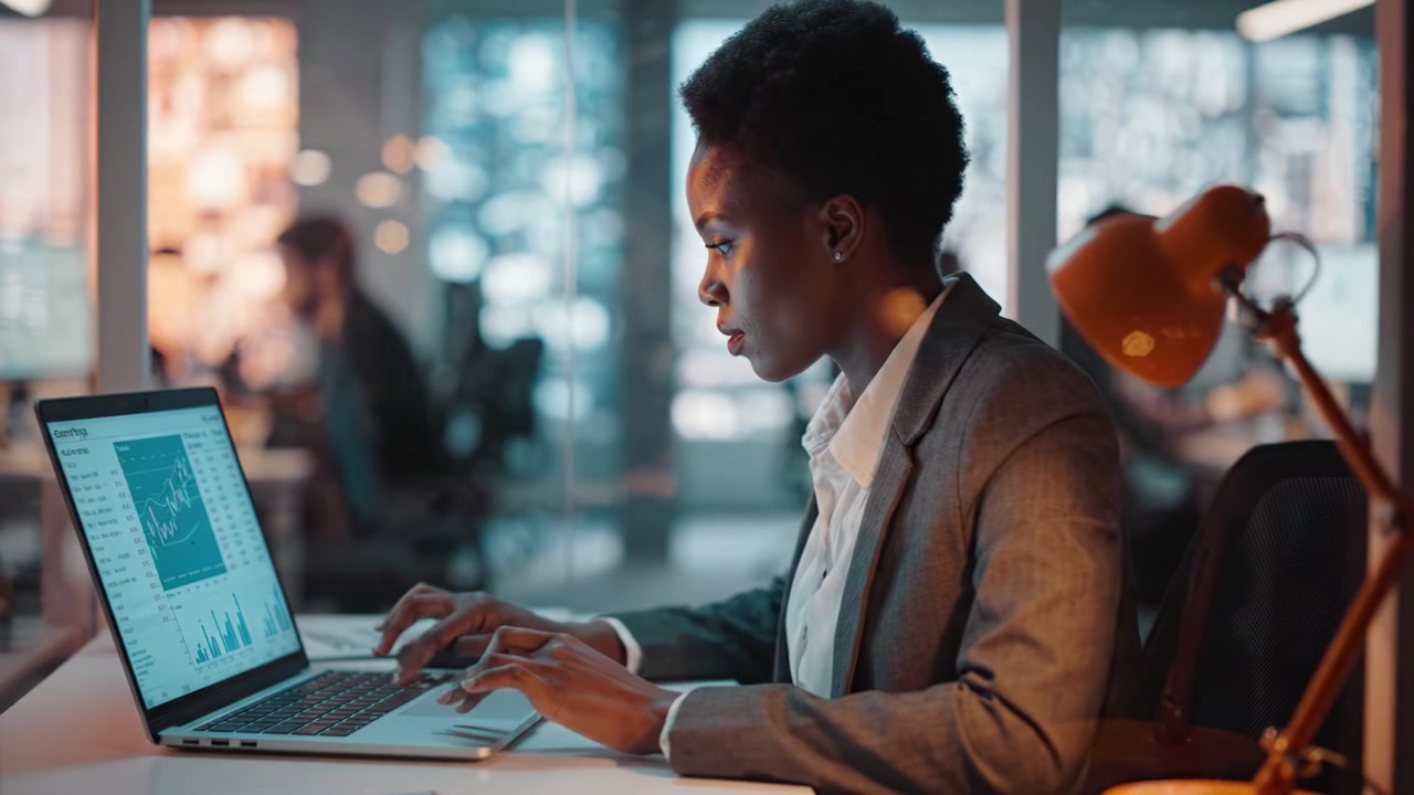 Focused Professional Working on Laptop with Data Analytics in Modern Office Environment, Surrounded by Colleagues and Soft Illuminated Workspace
