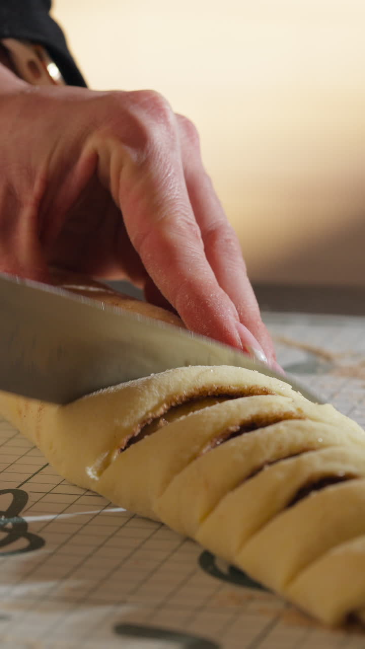 Close-up Of Hand Using Kitchen Knife In Slicing Log Of Rolled Dough For Cinnamon Rolls. vertical shot