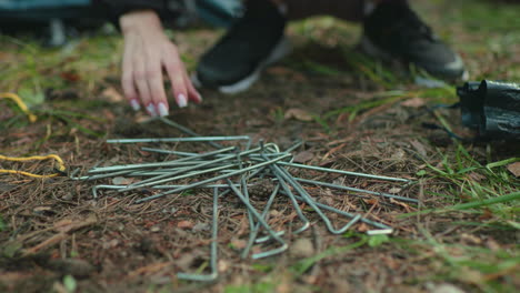 close up of tent stakes lying on pine covered ground while camper squats nearby reaching for equipment during setup in forest campsite surrounded by greenery and scattered tools