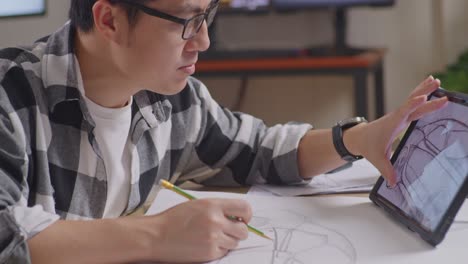 close up of asian male looking at a tablet and drawing on paper while working on a car design sketch on table in the studio