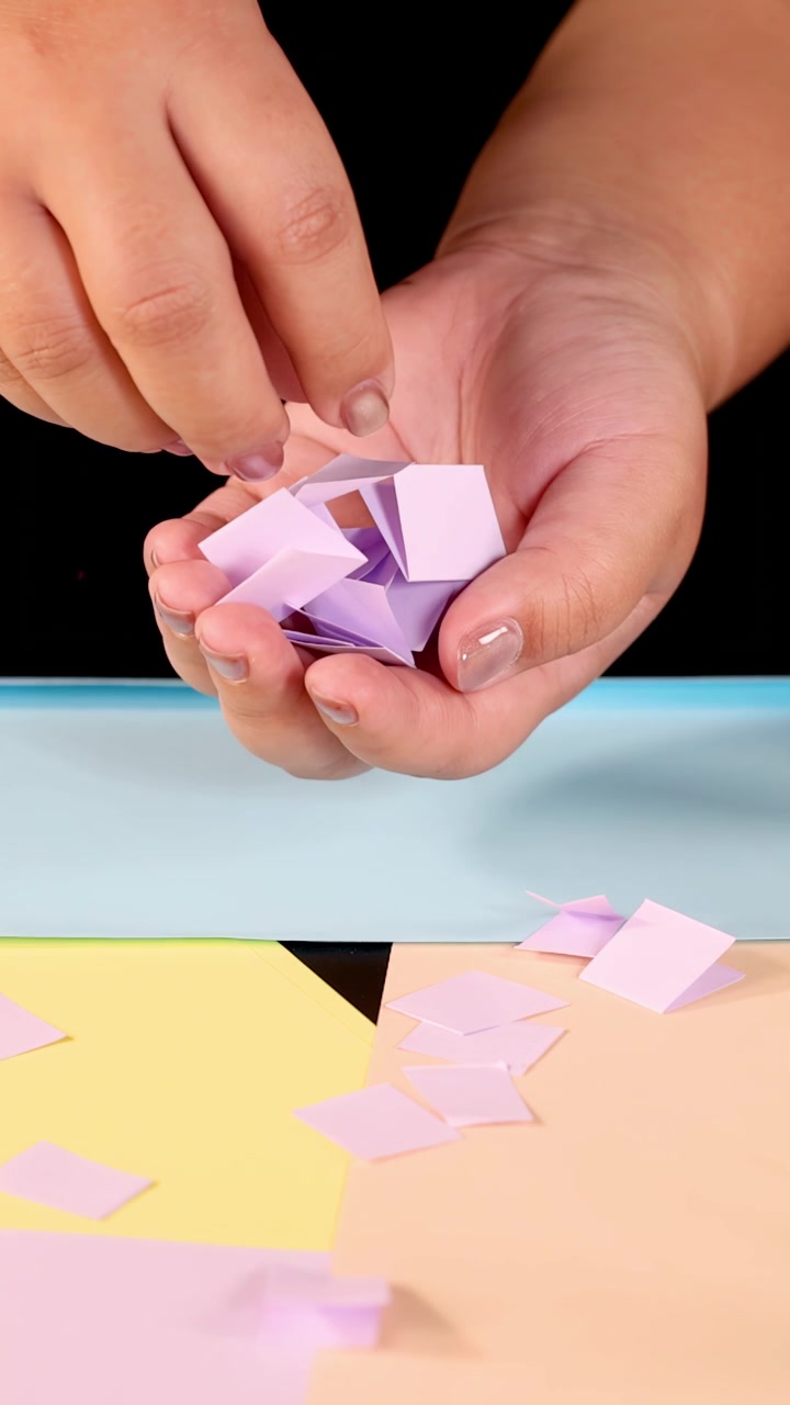 Mani che raccolgono quadrati di carta origami pastello da una superficie colorata sotto un'illuminazione da studio brillante e uniforme