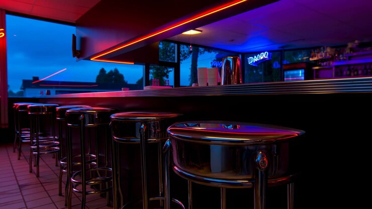 Sgabelli in cromo lucido fiancheggiano un lungo bancone da tavola calda da un punto di vista ravvicinato e basso, enfatizzando i sedili riflettenti, i telai tubolari, il piano del bancone scuro, l'insegna al neon rossa e un ambiente di ospitalità retrò pulito.