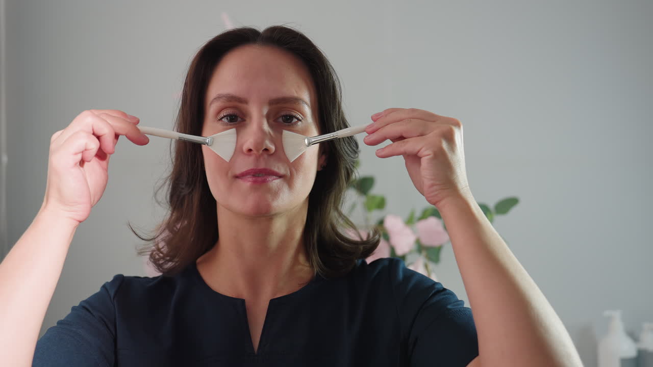 portrait view of cheerful female massage therapist holding two silicone facial brushes brushing cheeks with playful strokes under warm natural light in stylish spa treatment room with floral decor