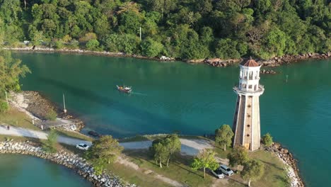 un dron de tiro cinematográfico vuela alrededor del faro del muelle de perdana, siguiendo un barco de pesca que navega por la vía fluvial hacia el puerto deportivo de telaga rodeado de hermosa naturaleza en la isla de langkawi, kedah, malasia