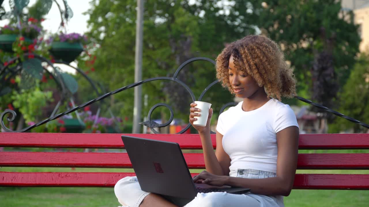 Beautiful young african american woman working on laptop on a bench in the city park and drinks coffee