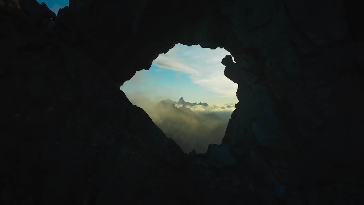 Aerial drone flying closer and through a dark colored silhouette like hole in a cliff or a mountains during sunset or sundown in Valle D'Aosta Italy. Through the opening blue sky with clouds visible.
