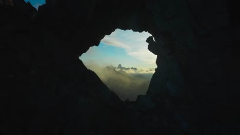 aerial drone flying closer and through a dark colored silhouette like hole in a cliff or a mountains during sunset or sundown in valle d'aosta italy. through the opening blue sky with clouds visible.