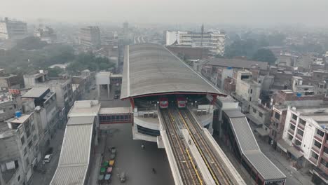 Aerial-drone-rotating-shot-over-orange-line-metro-trains-waiting-in-a-station-on-McLeod-Road,-Lahore,-Pakistan-during-morning-time