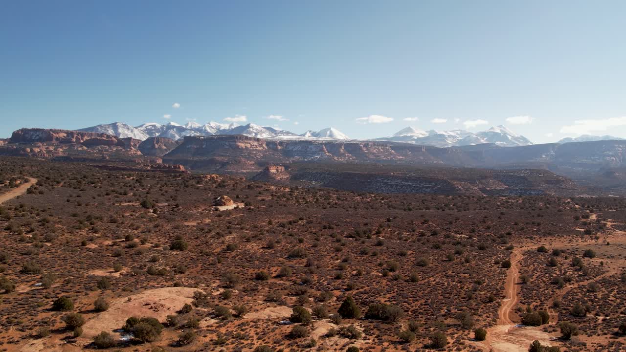 A high-flying drone shot over a remote dirt road cutting through the vast and unique desert land near Moab, Utah, with the snowy Rocky Mountains towering in the distance