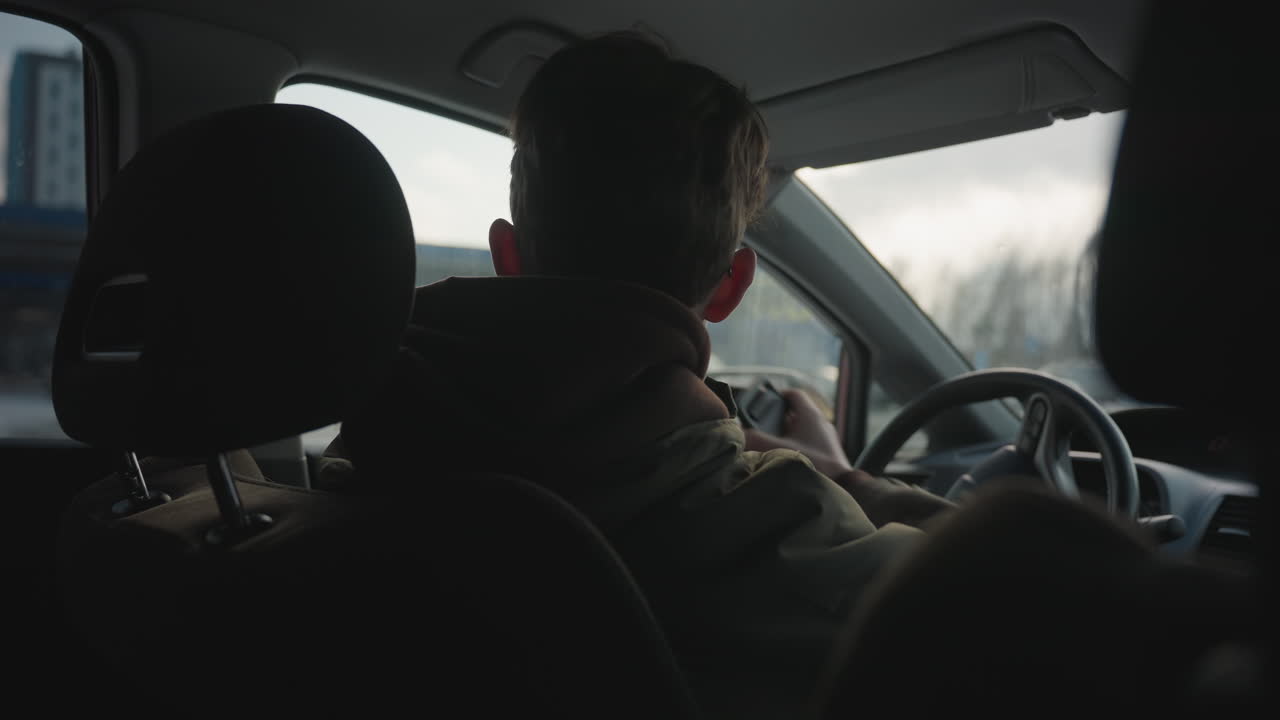 young boy inside parked car removes seatbelt while leaning forward, wearing glasses and hoodie under jacket, steering wheel in foreground, soft natural light entering window with car visible outside