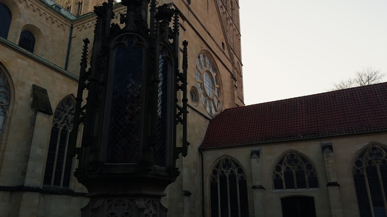 Struttura architettonica gotica nel cortile della Cattedrale di San Paolo a Münster, Germania. Facciata storica della chiesa con rosone e dettagli in pietra medievali sotto un cielo coperto.
